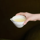 A hand holding a textured white ceramic pitcher filled with golden Toolong oolong tea liquor against a moss green background