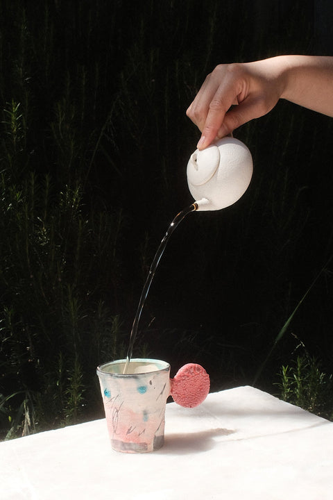 A hand pours dark liquid from a small white teapot into a clear patterned cup resting on a white tablecloth, with a pink sponge nearby and a dark outdoor background.