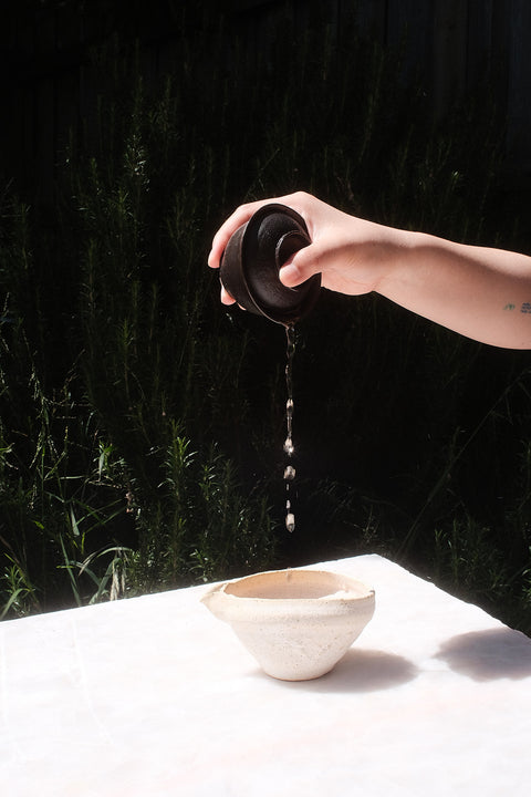 A hand holds a dark ceramic teapot lid over a white table, with water dripping into a small beige bowl. The background is a dark, shadowed outdoor setting with greenery.