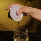 A hand holds a white ceramic gaiwan lid and pours tea into a clear glass pouring vessel on a dark table, with a warm yellow background.