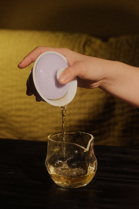 A hand holds a white ceramic gaiwan lid and pours tea into a clear glass pouring vessel on a dark table, with a warm yellow background.