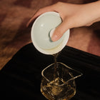 A hand holds a white ceramic gaiwan lid and pours tea into a clear glass pouring vessel on a dark table, with a warm brown textured background.