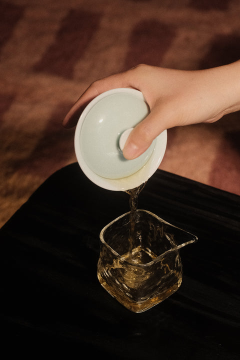 A hand holds a white ceramic gaiwan lid and pours tea into a clear glass pouring vessel on a dark table, with a warm brown textured background.