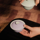 Two hands holding traditional Chinese gaiwan tea bowls, one with a pale lavender exterior and one with a pale mint green exterior, against a dark background.