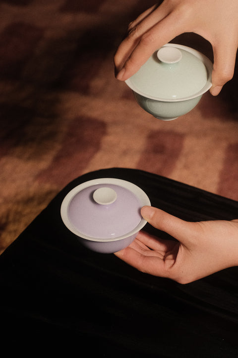 Two hands holding traditional Chinese gaiwan tea bowls, one with a pale lavender exterior and one with a pale mint green exterior, against a dark background.