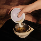 A hand pouring tea from a white ceramic gaiwan with a pale purple lid into a metal and ceramic tea infuser basket over a dark surface.