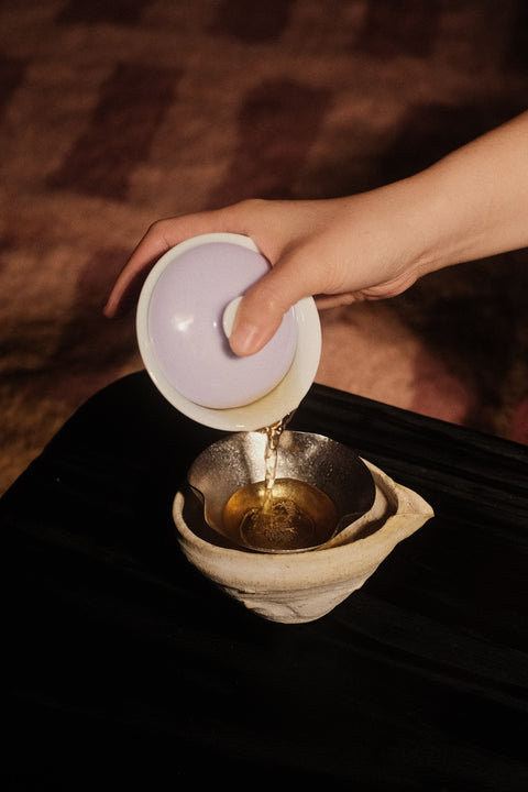 A hand pouring tea from a white ceramic gaiwan with a pale purple lid into a metal and ceramic tea infuser basket over a dark surface.