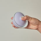 A hand holding a small round lavender and white container with a matching lid against a plain light gray background.