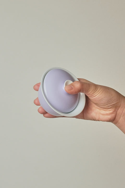 A hand holding a small round lavender and white container with a matching lid against a plain light gray background.