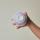 A hand holding the base of a small, pale lavender ceramic bowl against a plain, warm beige background.