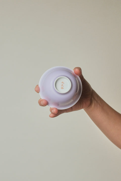 A hand holding the base of a small, pale lavender ceramic bowl against a plain, warm beige background.