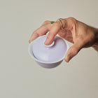 A hand holding a small white ceramic lidded bowl with a pale lavender-purple interior against a solid light gray background.