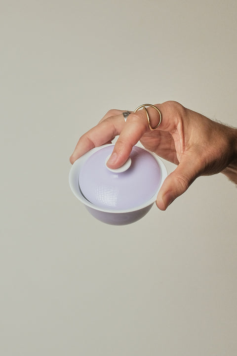 A hand holding a small white ceramic lidded bowl with a pale lavender-purple interior against a solid light gray background.