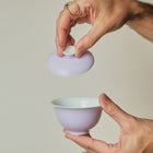 A person's hands holding a light purple ceramic tea bowl and its matching lid against a solid light beige background.