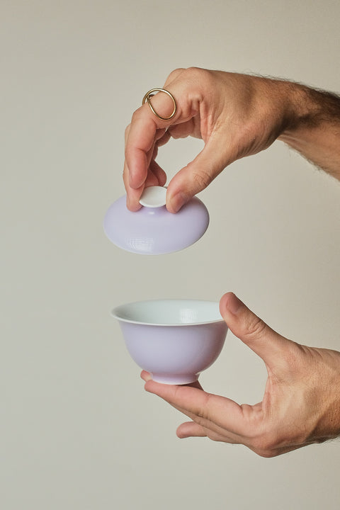 A person's hands holding a light purple ceramic tea bowl and its matching lid against a solid light beige background.