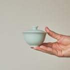 A hand holding a small pale celadon-green ceramic lidded bowl against a plain warm beige background.