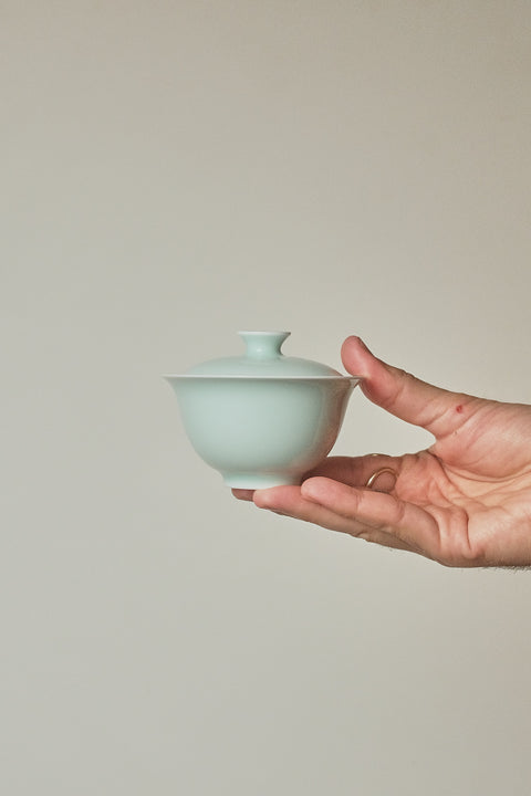 A hand holding a small pale celadon-green ceramic lidded bowl against a plain warm beige background.