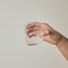 A hand holding a small clear glass tumbler against a plain light gray background.