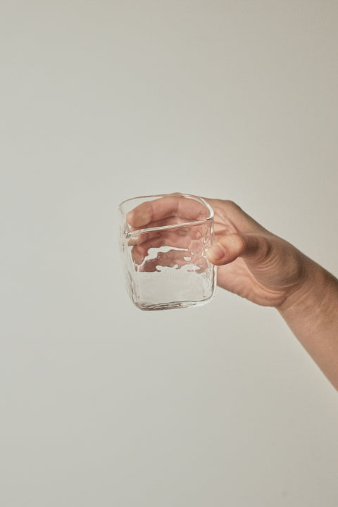 A hand holding a small clear glass tumbler against a plain light gray background.