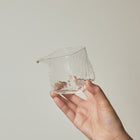 A hand holding a small, clear, hand-blown glass cup with a textured, hammered surface against a plain, warm off-white background.
