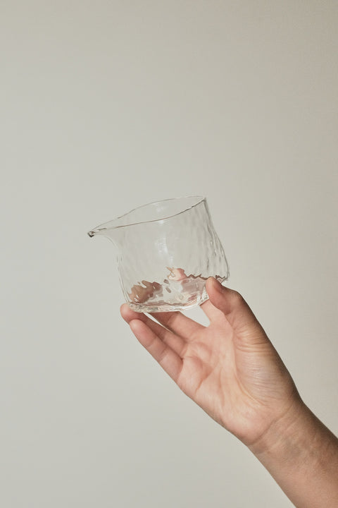 A hand holding a small, clear, hand-blown glass cup with a textured, hammered surface against a plain, warm off-white background.