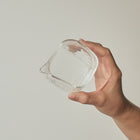 A hand holding a small clear glass container with a square shape and a small pouring spout, set against a plain light gray background.