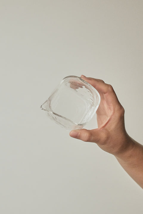 A hand holding a small clear glass container with a square shape and a small pouring spout, set against a plain light gray background.