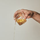 A hand holding a small clear glass container filled with amber liquid, with the liquid pouring in a thin stream against a plain light beige background.
