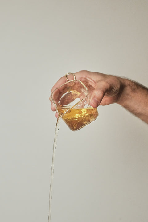 A hand holding a small clear glass container filled with amber liquid, with the liquid pouring in a thin stream against a plain light beige background.