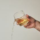 A hand holding a clear textured glass tumbler with amber liquid, from which a stream of liquid is pouring downward, against a plain light gray background.