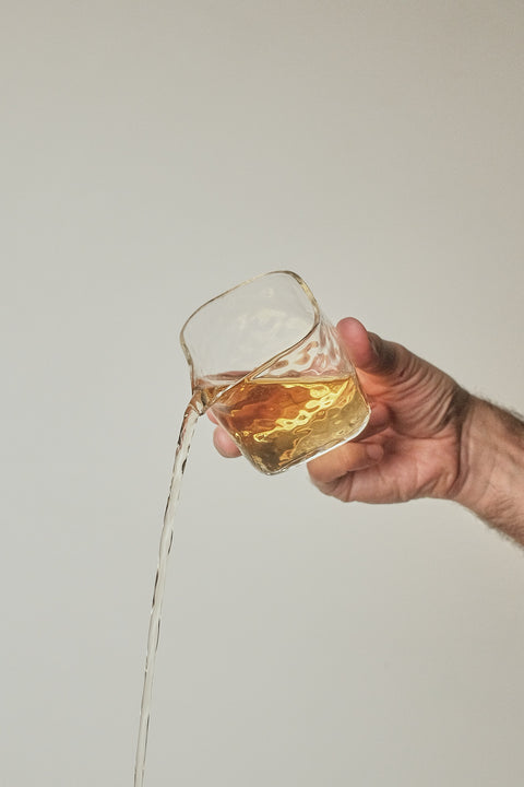 A hand holding a clear textured glass tumbler with amber liquid, from which a stream of liquid is pouring downward, against a plain light gray background.