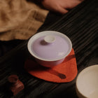 A ceramic tea set with a pale lavender-purple lid and white body, resting on a textured orange coaster on a dark wooden table. A person's hand and clothing are visible in the softly blurred background.