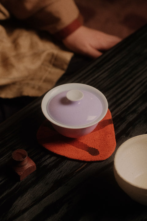 A ceramic tea set with a pale lavender-purple lid and white body, resting on a textured orange coaster on a dark wooden table. A person's hand and clothing are visible in the softly blurred background.