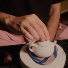 A close-up of a person's hand resting on the lid of a small matte white ceramic teapot, which sits on a round textured base with a blue patterned cloth underneath.