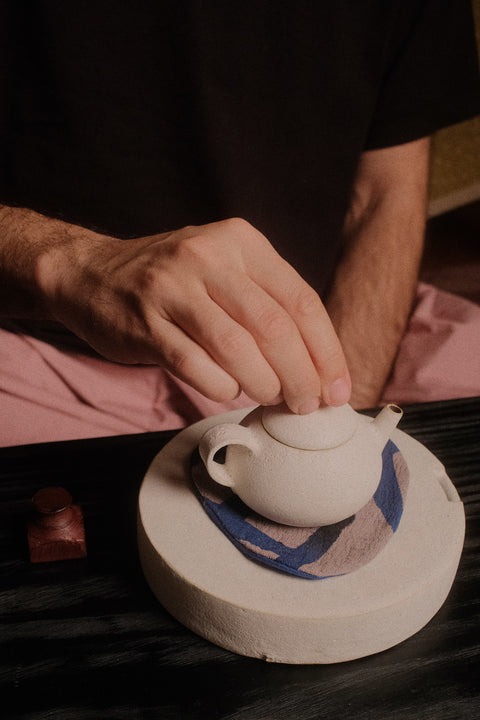A close-up of a person's hand resting on the lid of a small matte white ceramic teapot, which sits on a round textured base with a blue patterned cloth underneath.