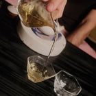 A hand pouring a clear amber liquid from a faceted glass pouring vessel into two small clear drinking glasses on a dark wooden table.