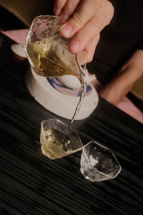 A hand pouring a clear amber liquid from a faceted glass pouring vessel into two small clear drinking glasses on a dark wooden table.