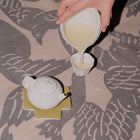 A hand pouring light-colored tea from a white ceramic pitcher into a small white cup, with a white teapot resting on a yellow coaster on a gray and beige patterned fabric surface.