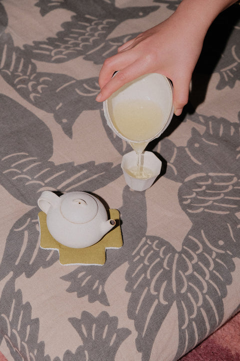 A hand pouring light-colored tea from a white ceramic pitcher into a small white cup, with a white teapot resting on a yellow coaster on a gray and beige patterned fabric surface.
