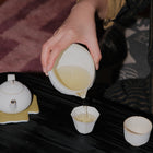 A hand pouring light yellow tea from a white ceramic teapot into small cups on a dark table, with a teapot and cup also visible.