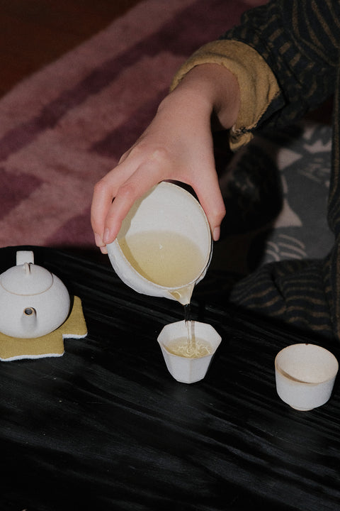 A hand pouring light yellow tea from a white ceramic teapot into small cups on a dark table, with a teapot and cup also visible.