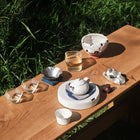 A ceramic tea set arranged on a wooden table outdoors, with grass visible in the background. The set includes a white teapot, matching cups, a bowl, and small glass vessels under bright natural sunlight.