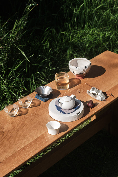 A ceramic tea set arranged on a wooden table outdoors, with grass visible in the background. The set includes a white teapot, matching cups, a bowl, and small glass vessels under bright natural sunlight.