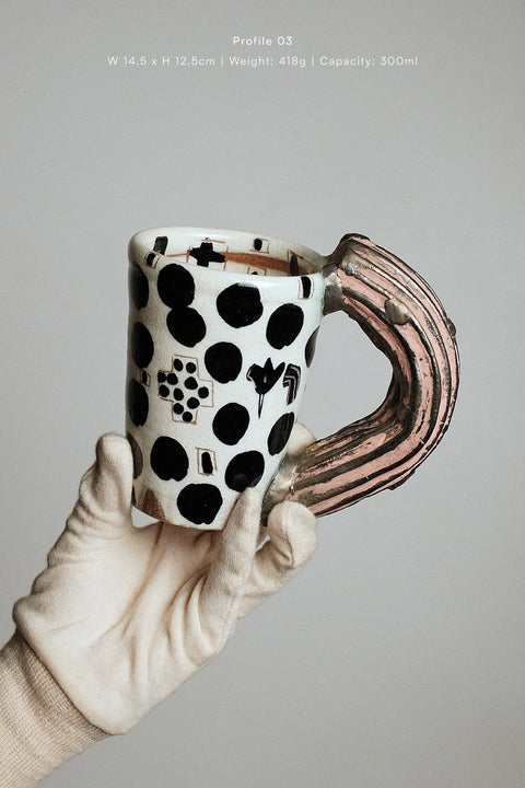 A hand wearing a cream-colored glove holds a ceramic mug with a white base, black polka dots, and a curved handle featuring a pink and brown striped pattern. The background is a solid light gray, and text at the top displays product specifications.