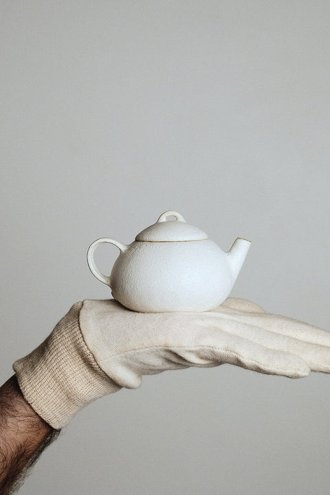 White ceramic teapot held by a gloved hand against a plain background
